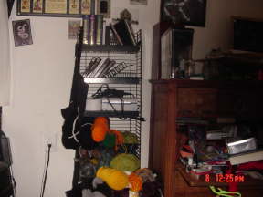 A picture of a cluttered, metal shelf & old, wooden bureau. On the shelf there are three Antiquarian Sticker Books, a Goosebumps book, a couple of X-Files books, The Hitchhiker's Guide to The galaxy, The Devil's Dictionary, & multiple journals of various shapes. There is also plenty of yarn for making projects, & a thermal printer for selling those projects. A home-crocheted black bag with a white X is hanging from the shelf. On the wall there are multiple tarot posters/hangings, clippings from magazines featuring old New England architecture/industry, a calendar, & paintings from a local artist. A metal detector leans against the wall. The bureau is completely covered in crafting supplies, toys, nail polish, Brita water filter, & Altoids tins. A hyper-realistic baby hippo statue lurks in the shadows.